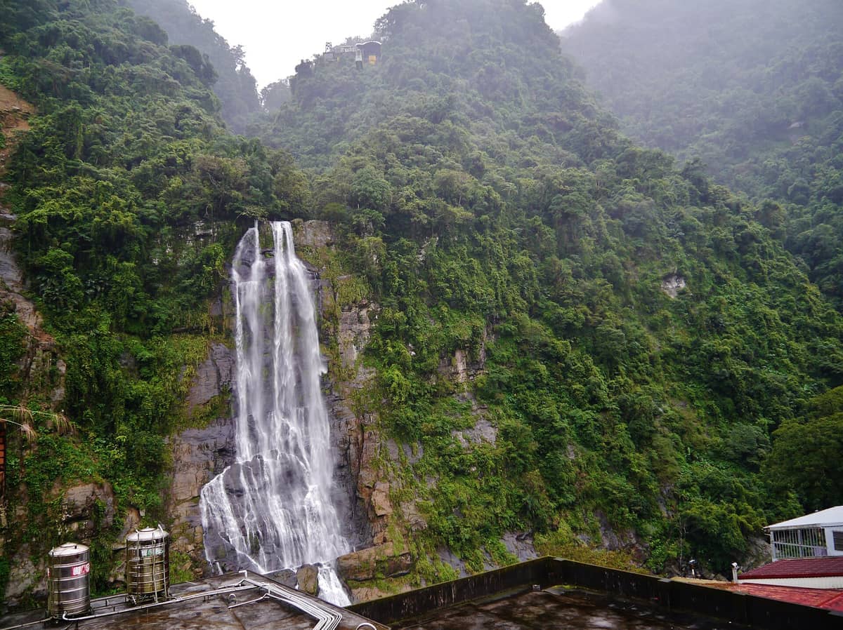 Der Wulai-Wasserfall, der eine grüne, bewaldete Klippe hinabstürzt, mit nebligen Bergen und Dächern im Vordergrund unter bedecktem Himmel.