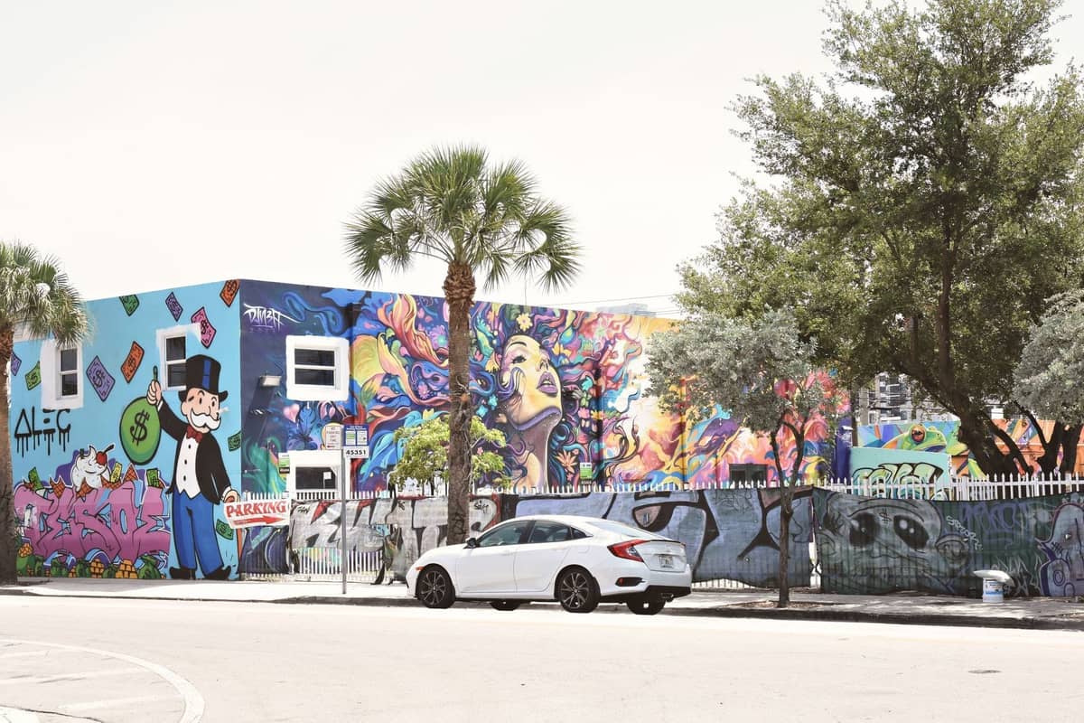 Brightly colored graffiti murals cover a building, palm trees out front, and a white car parked on the street in Wynwood, Miami.