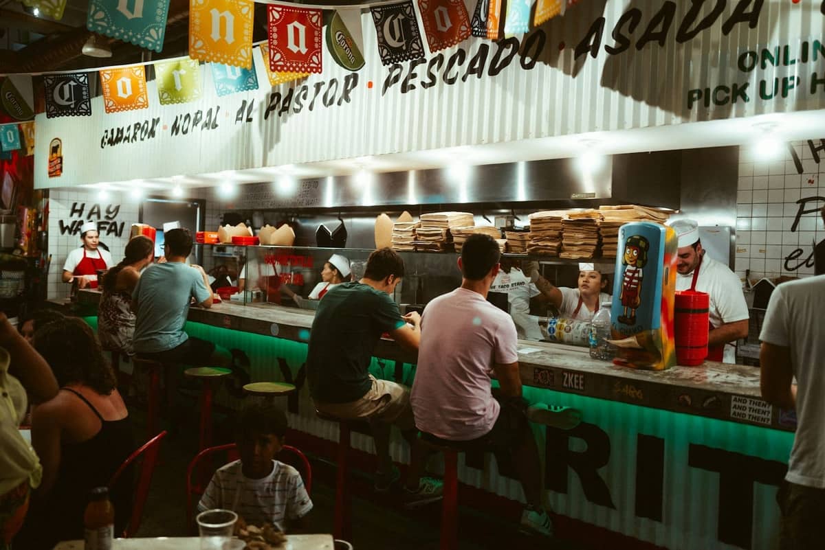 People seated at a lively taco bar with cooks preparing food behind the counter, festive decorations and menu signs above.