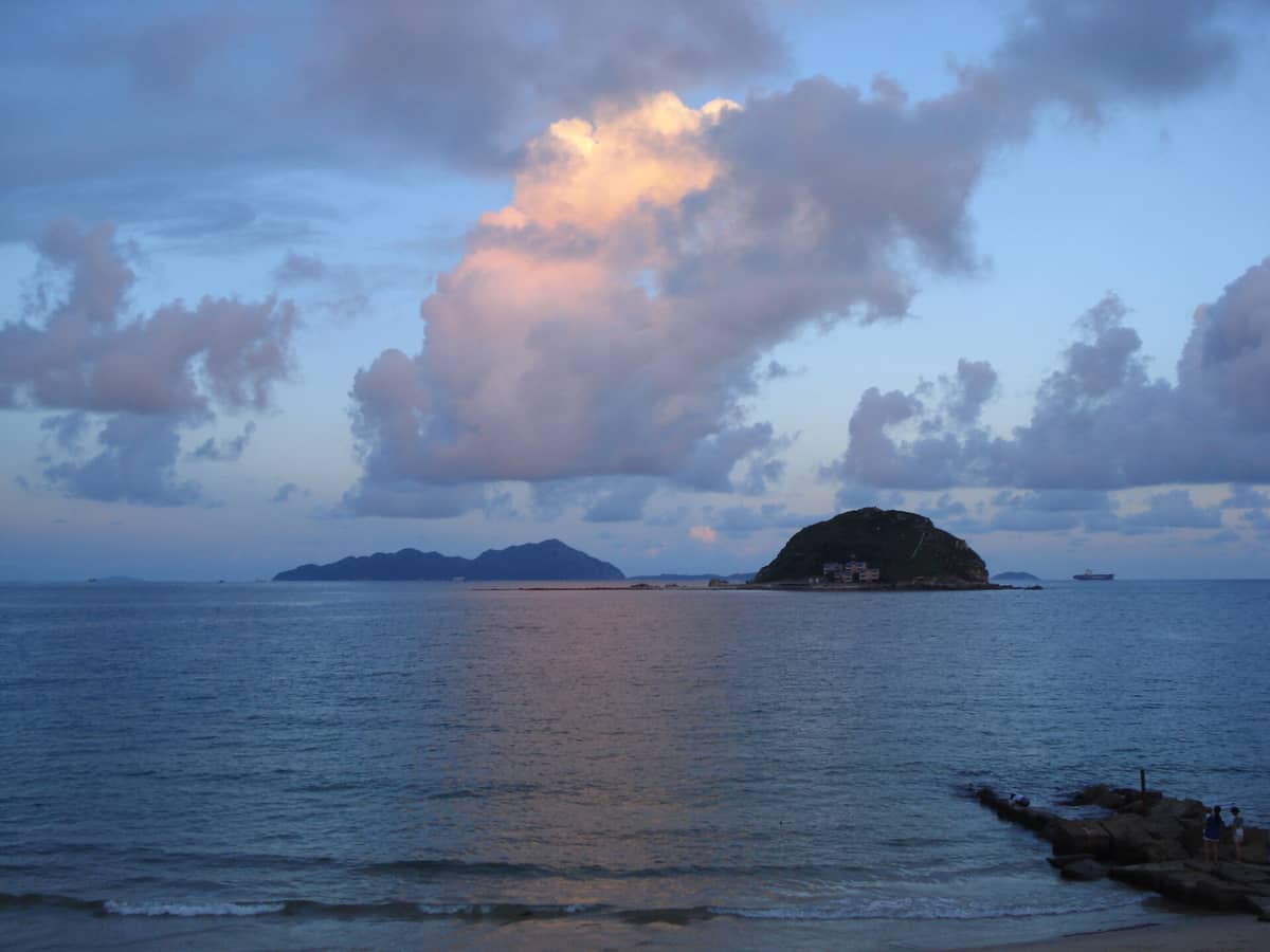 Vista da praia de Xichong ao entardecer, com águas azuis calmas, ilhas rochosas, nuvens dramáticas ao anoitecer e um distante promontório na península de Dapeng.