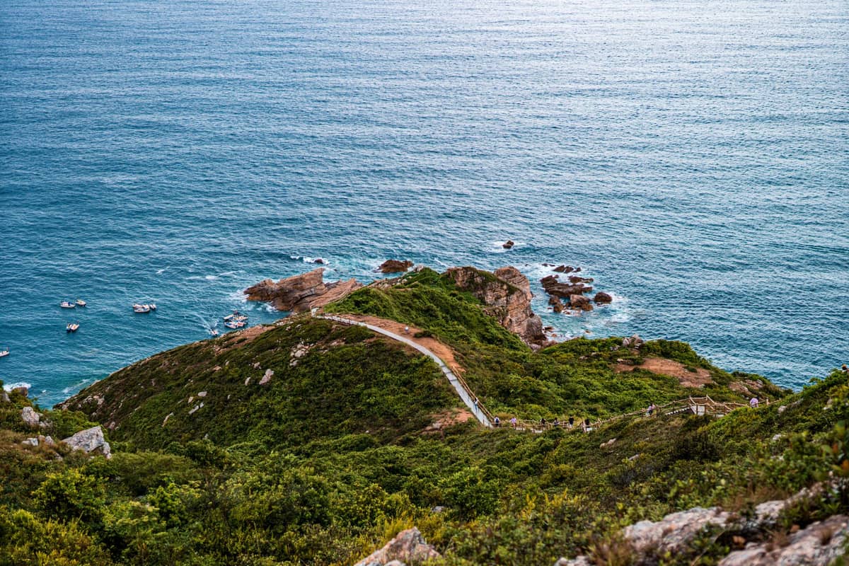 Vista panorâmica da trilha costeira Xichong–Dongchong sinuando pela beira de uma falésia verde com caminhantes, mar azul cristalino lá embaixo e afloramentos rochosos ao fundo.