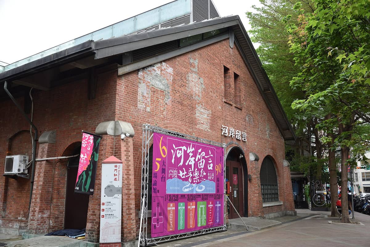 Die Außenansicht des Ximen Red House in Taipeh — ein historisches Backsteingebäude mit Schildern und Bannern am Eingang und Bäumen an der Seite.