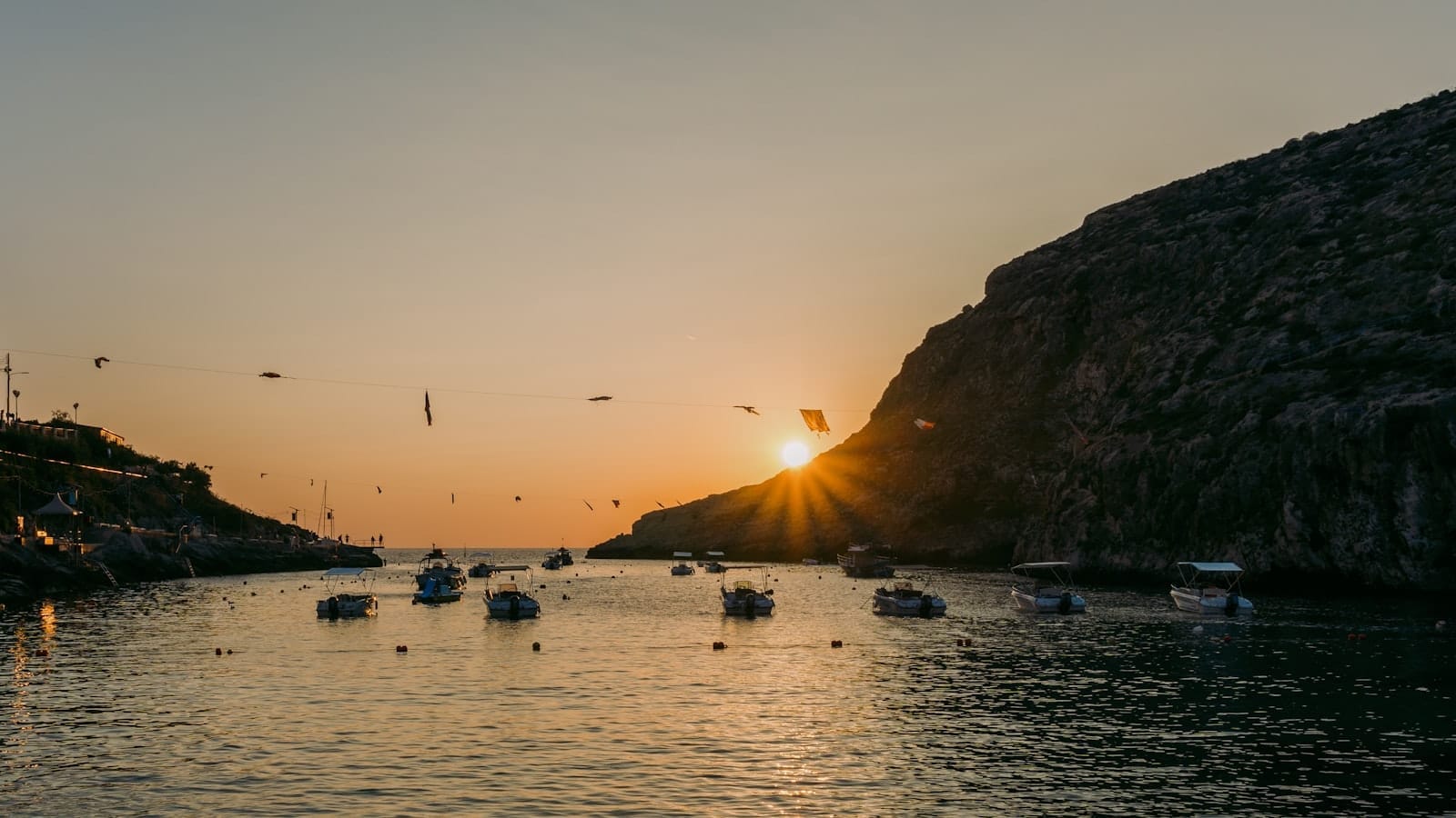 Tramonto sulla baia di Xlendi con acque turchesi calme, piccole barche e drammatiche scogliere calcaree che incorniciano la stretta insenatura.