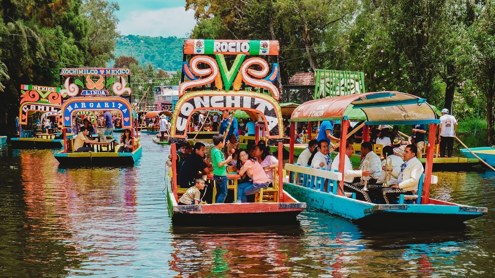 Trajineras dai colori vivaci piene di persone galleggiano lungo i Canali di Xochimilco, circondate da alberi verdi e vivaci riflessi sull'acqua.