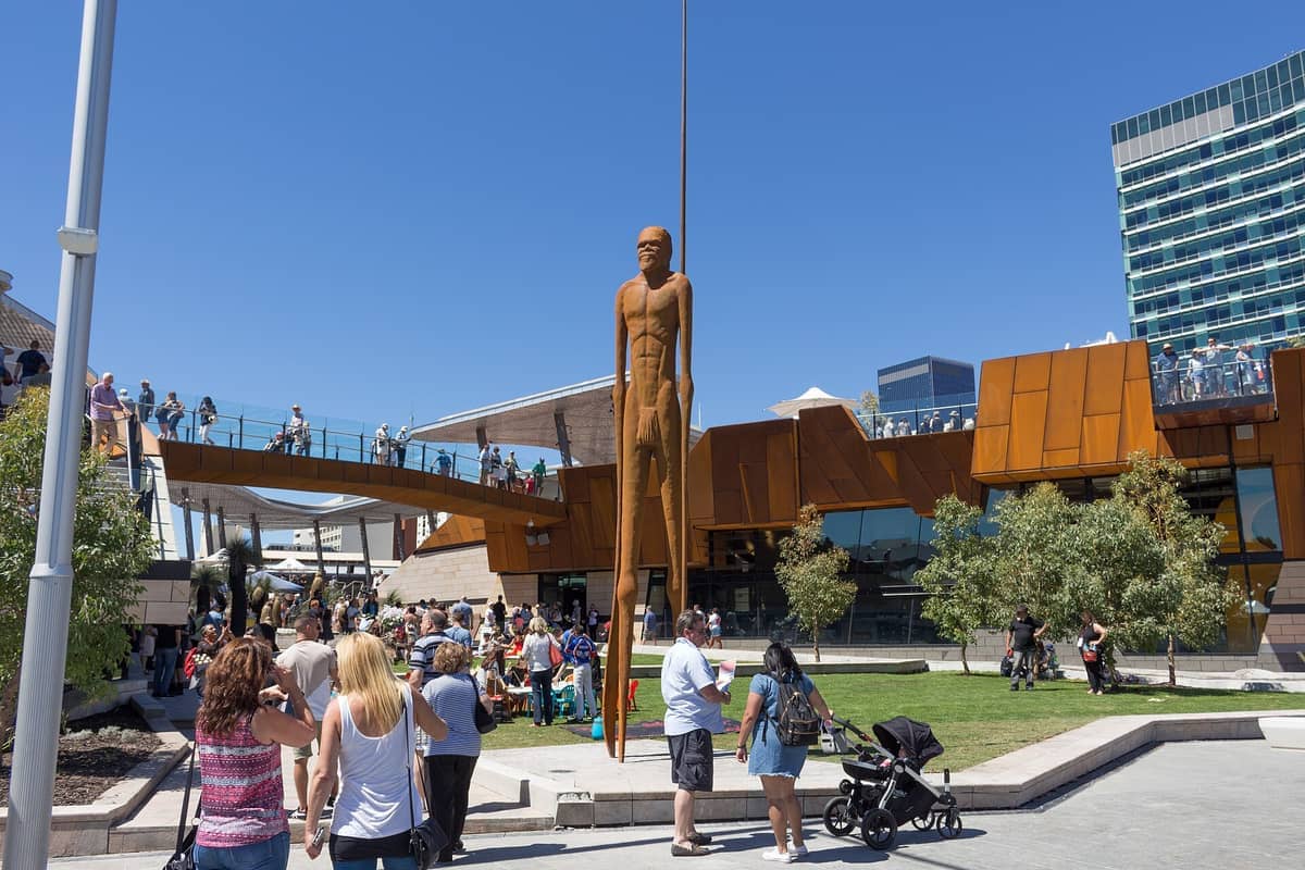 Multitud de personas disfrutando de un día soleado en Yagan Square, con la distintiva estatua aborigen alta y la arquitectura moderna en el centro de la ciudad de Perth.