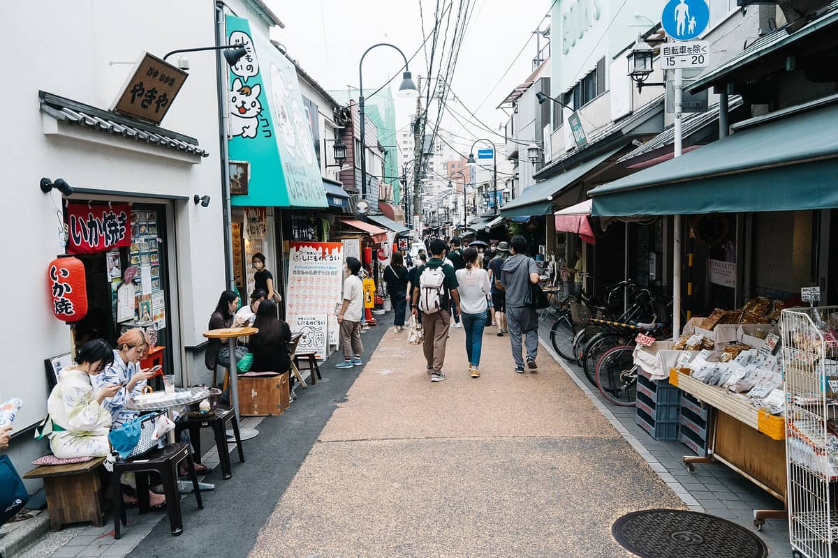 View along Yanaka Ginza shopping street in Tokyo showing local shops, street food stalls, and people walking and sitting outdoors.