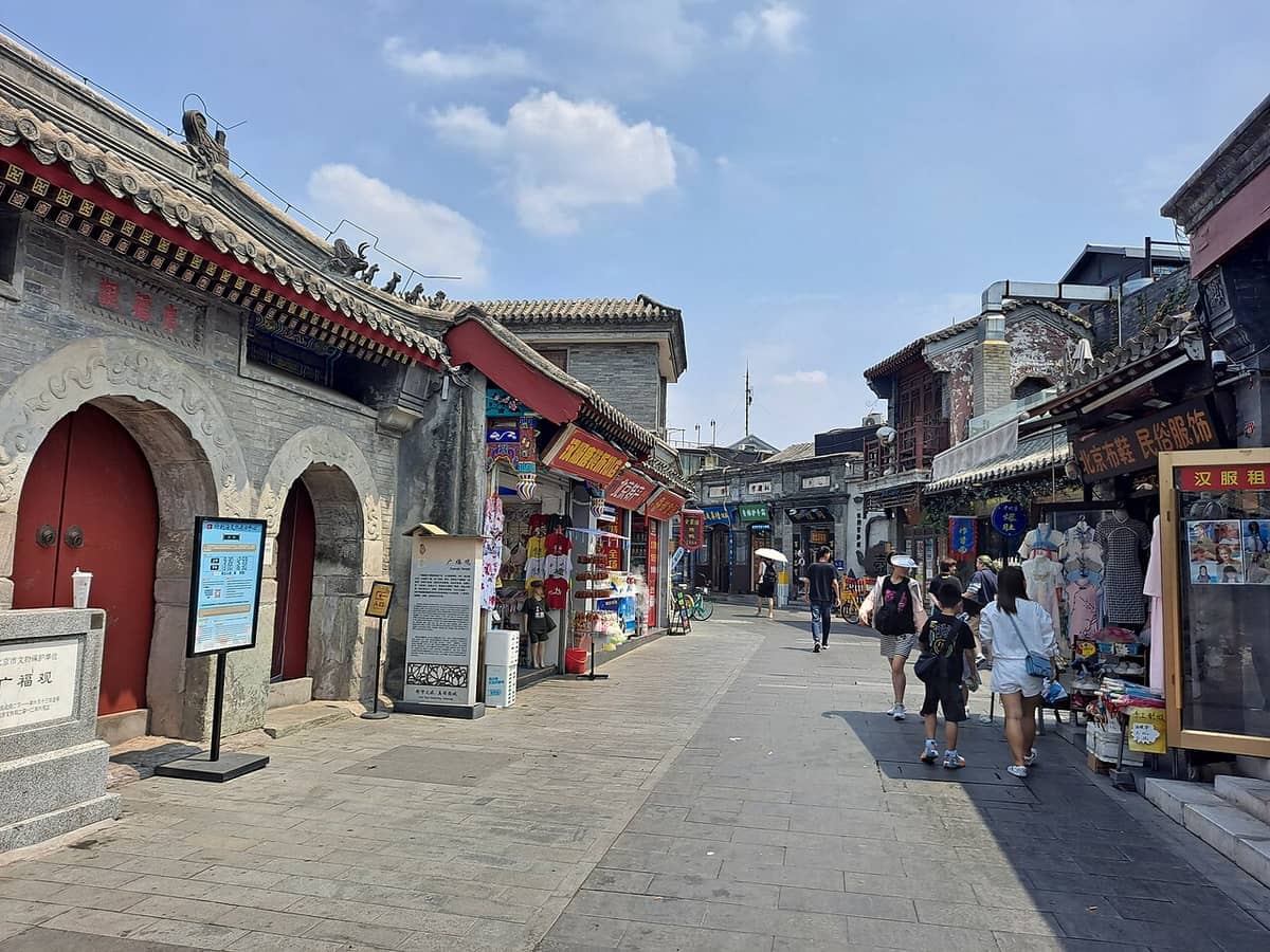 Die Yandaixie Street in Peking an einem klaren Tag: ein traditionelles graues Hofportal, Ladenfronten und Fußgänger auf dem gepflasterten Hutong.