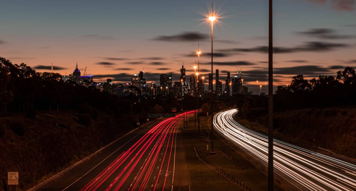 Movimentada autoestrada de múltiplas faixas ao anoitecer com rastros de luzes de carros em direção ao horizonte de Melbourne ao fundo.