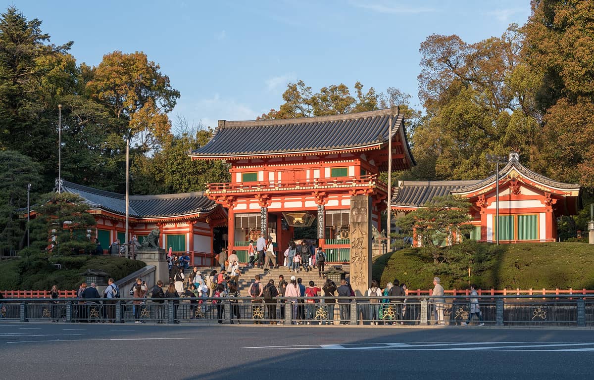 Crowds gather at the iconic two-story vermillion entrance gate of Yasaka Shrine in Kyoto, surrounded by lush greenery and traditional architecture on a sunny day.