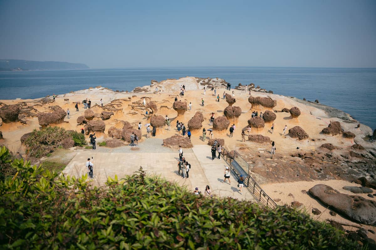 Weitblick auf die markanten pilzförmigen Felsformationen des Yehliu Geoparks an der Küste, mit Besuchern darunter und strahlend blauem Himmel.