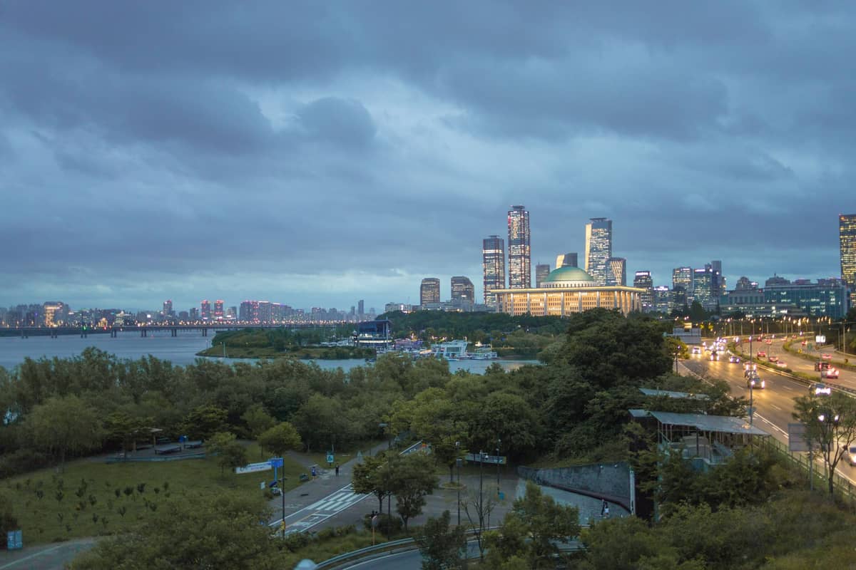 Vista do entardecer da ilha Yeouido com o prédio da Assembleia Nacional e sua cúpula verde, torres comerciais e o Rio Han em primeiro plano.