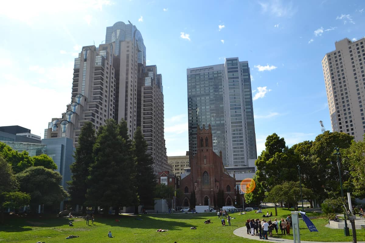 Yerba Buena Gardens a San Francisco, con persone che si rilassano sul prato verde, circondato da alberi e grattacieli moderni tra cui il Marriott Marquis e la storica chiesa di St. Patrick.