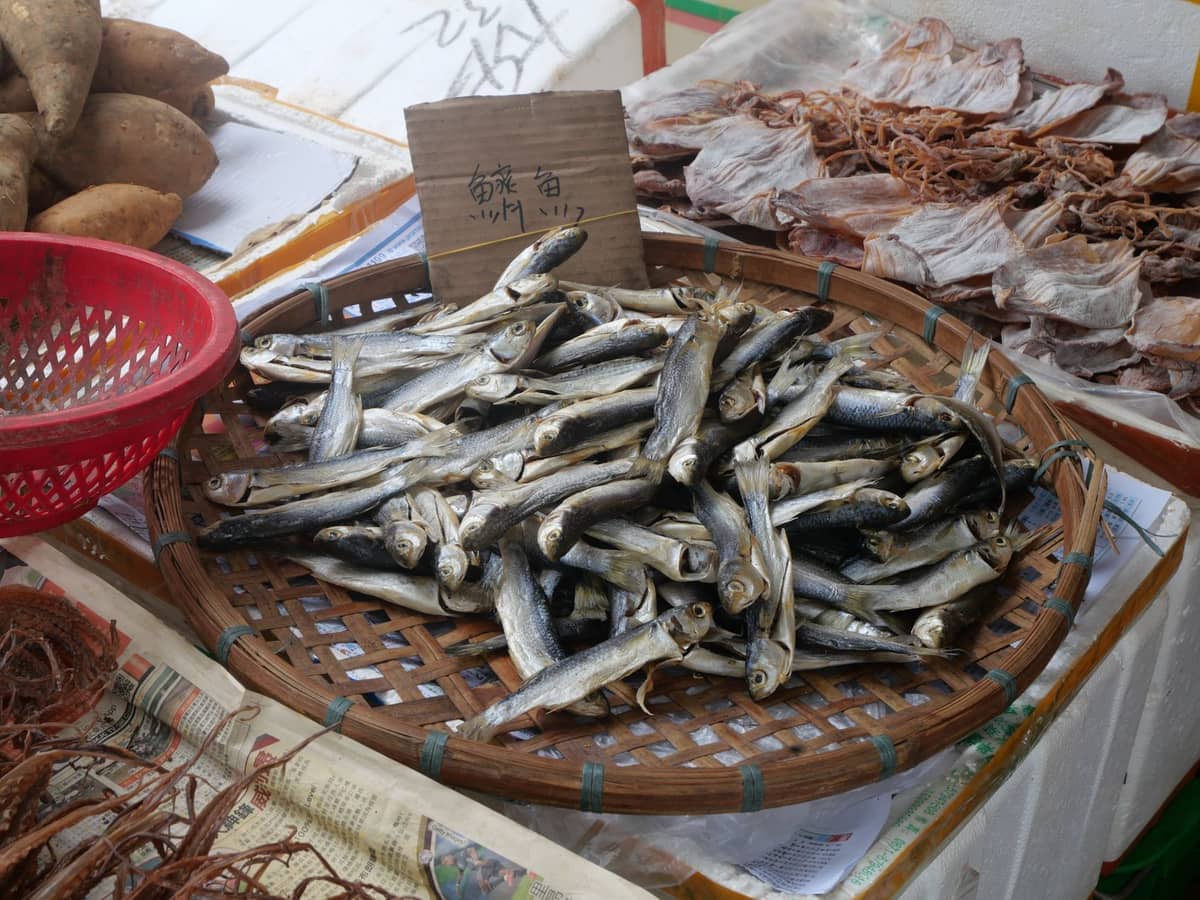 A woven basket filled with dried fish at a Yide Road Dried Seafood Street market stall, with other seafood and root vegetables nearby.