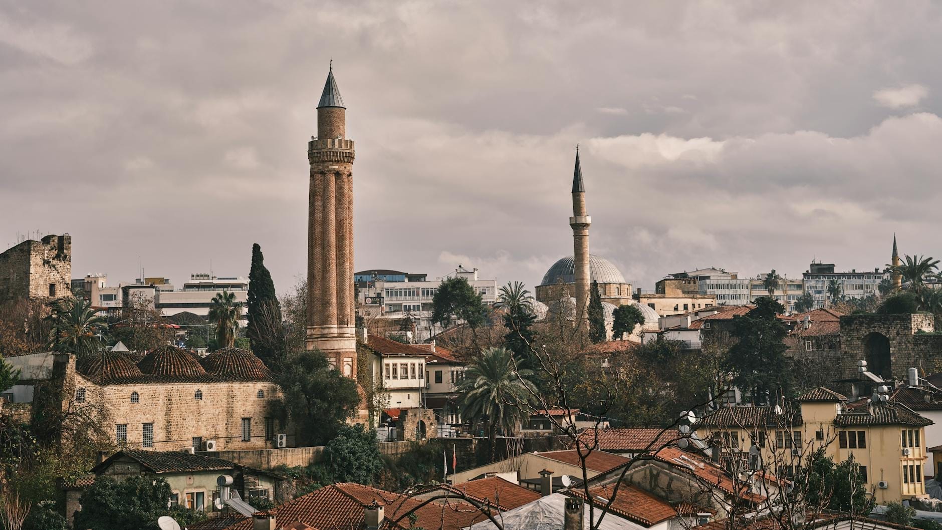 Vista panoramica del Minareto Scanalato che si erge sopra i tetti della città vecchia di Antalya sotto un cielo nuvoloso, circondato da edifici storici e rigogliosa vegetazione.