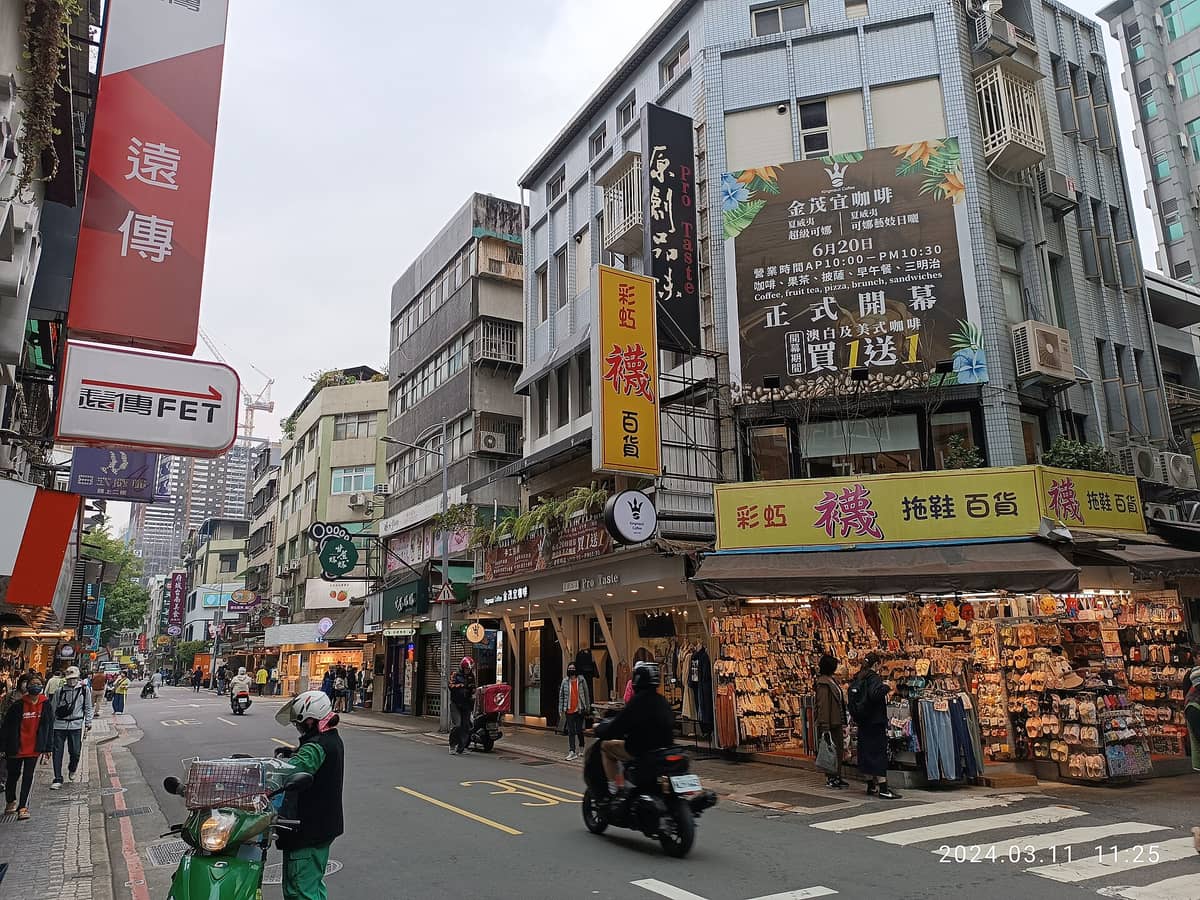 Belebte Tagesansicht der Yongkang Street in Taipeh mit Läden, Restaurants, Rollern und Fußgängern unter bunten Ladenschildern.