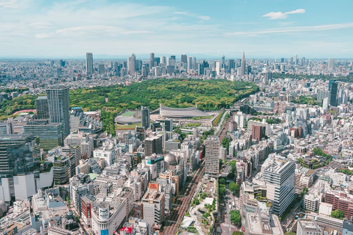 Aerial view of Yoyogi Park’s large green expanse surrounded by the Tokyo cityscape and skyscrapers under a bright, clear sky.