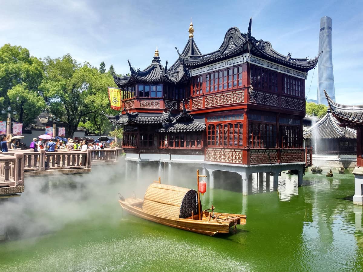 Traditioneller chinesischer Pavillon des Yu Gardens, im grünen Teich gespiegelt, mit einem Holzboot, Nebel, Touristen und einem modernen Wolkenkratzer im Hintergrund, Shanghai.