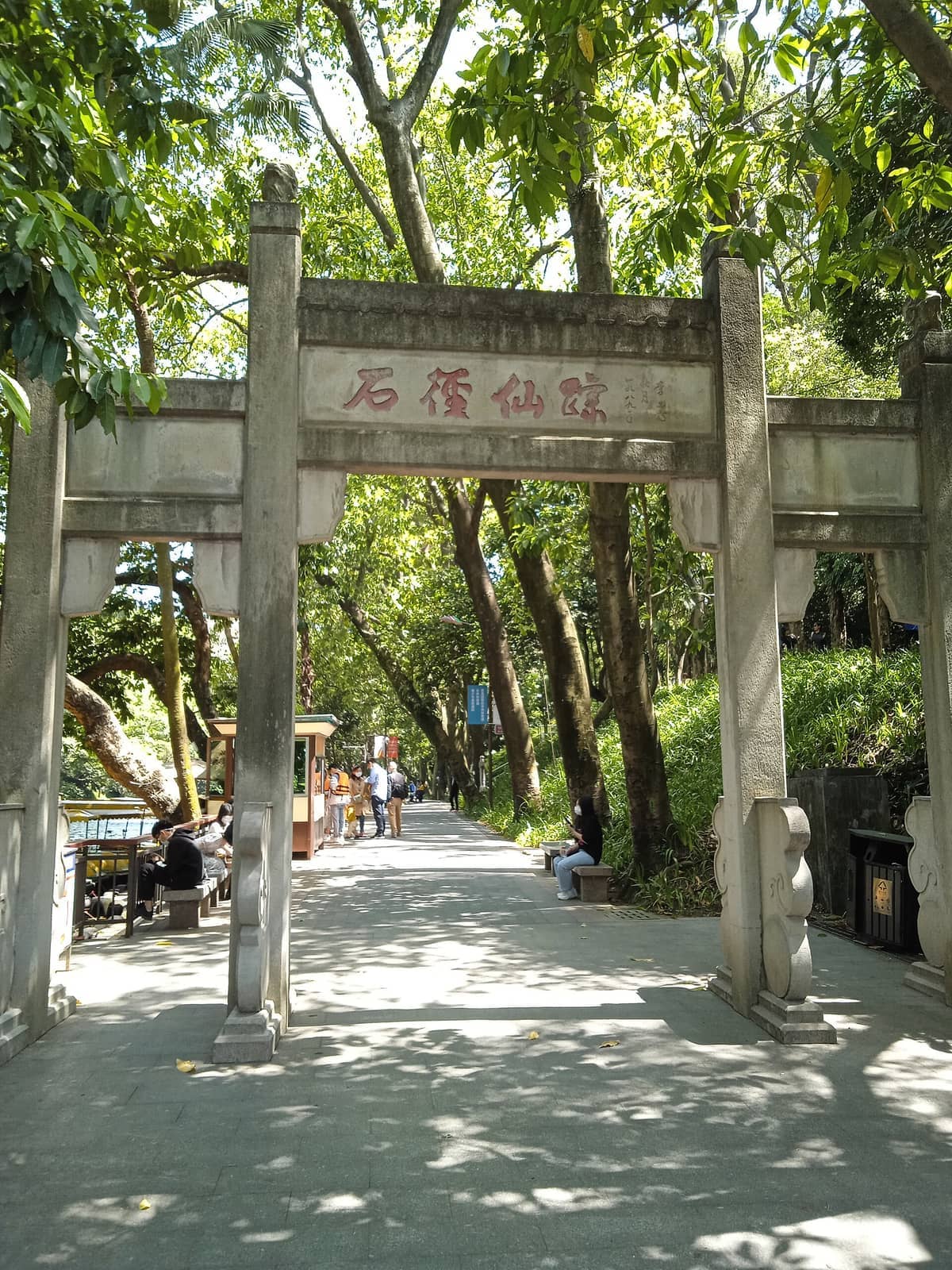 Stone archway entrance surrounded by lush greenery and tree-lined path in Yuexiu Park, with visitors sitting and strolling in daylight.