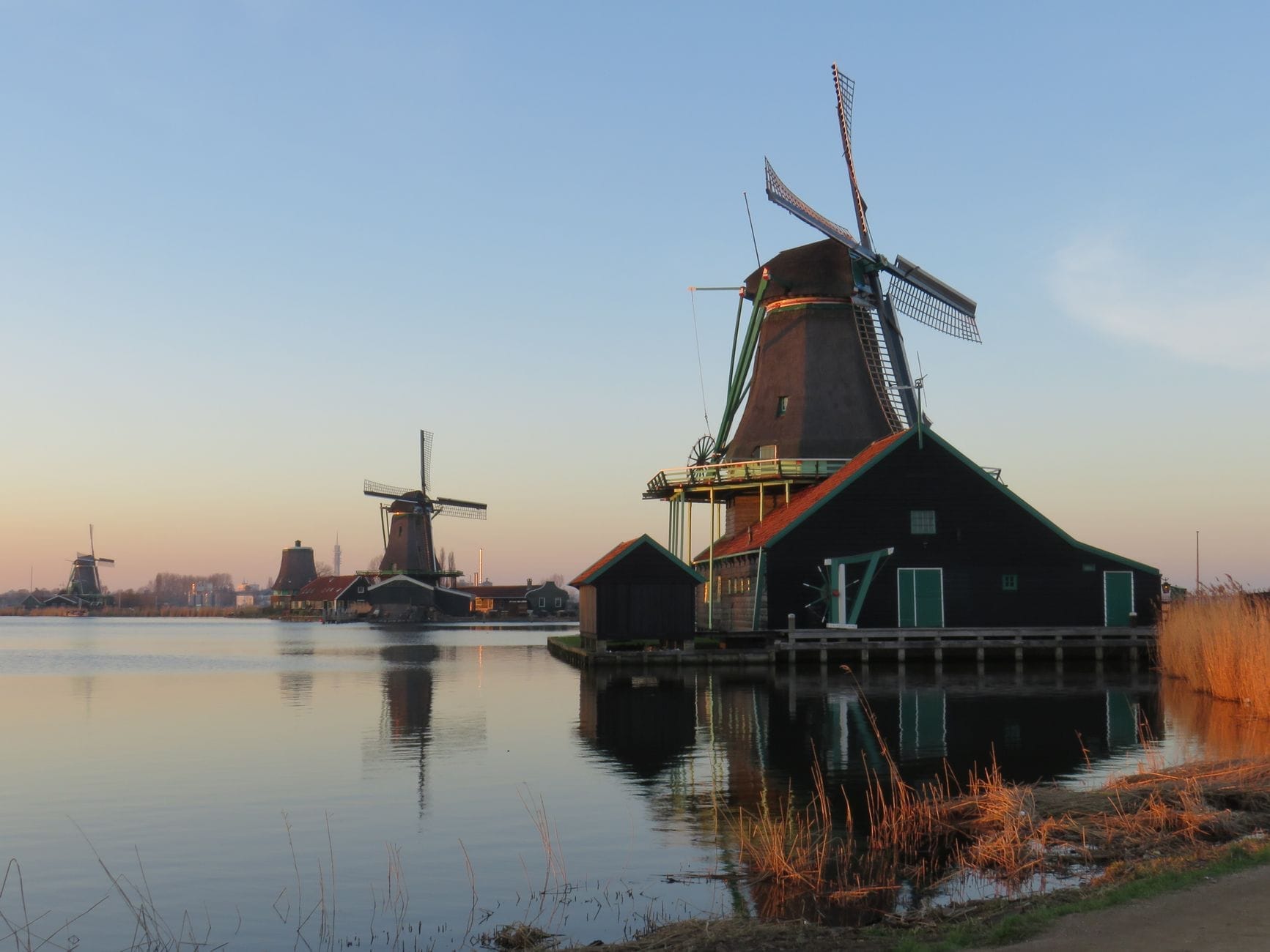 Clásicos molinos de viento holandeses y casas de madera verde reflejados en el agua tranquila del pueblo de Zaanse Schans durante un sereno amanecer o atardecer.