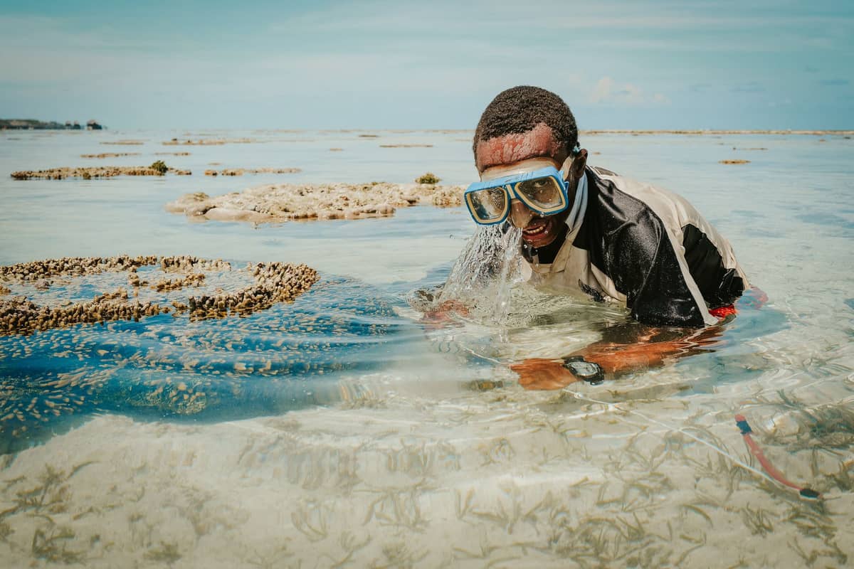 Snorkelista w okularach nurkowych w płytkiej, przejrzystej wodzie wśród kolonii koralowców na tropikalnej rafie; w tle błękitne niebo i odsłonięte koralowce podczas odpływu.