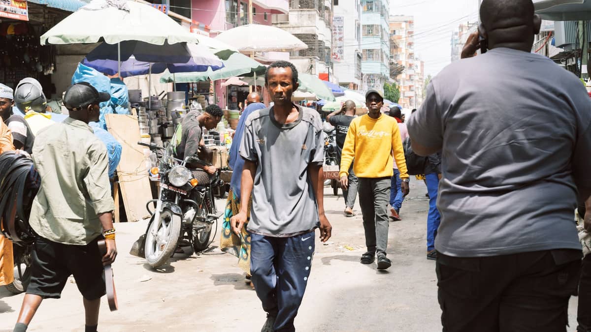 Animada escena de calle comercial con actividad de mercado, personas caminando y tiendas en Dar es Salaam bajo sombrillas y edificios de la ciudad.