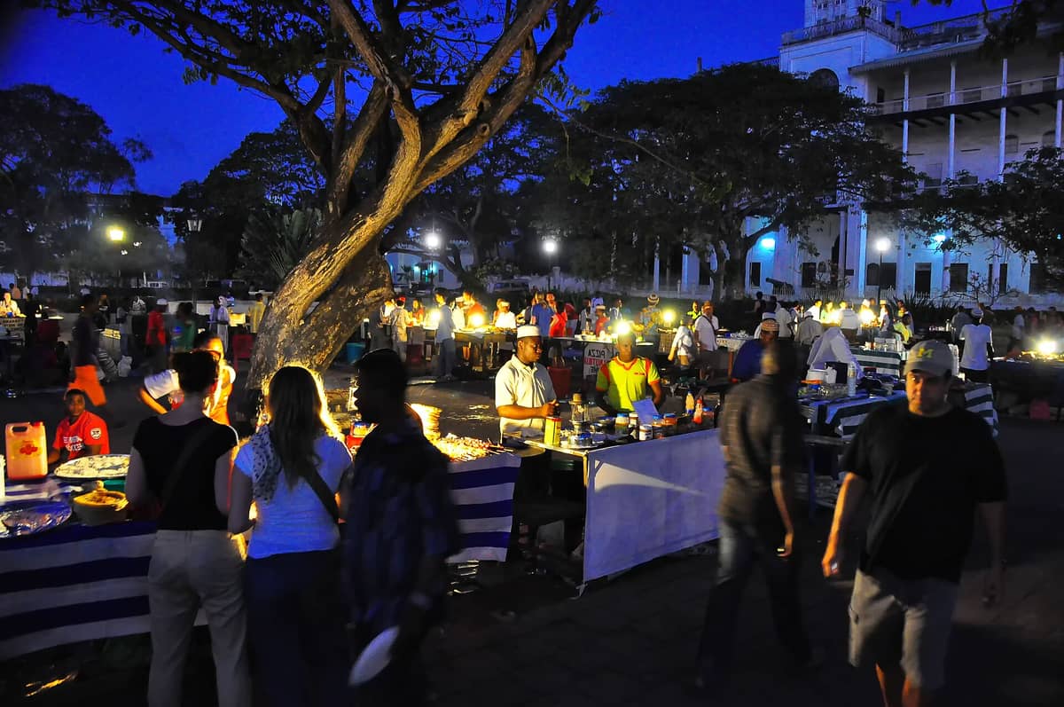 Forodhani Gardens night market at dusk with food stalls, vendors, and visitors illuminated by lanterns and surrounded by large trees and historic buildings.