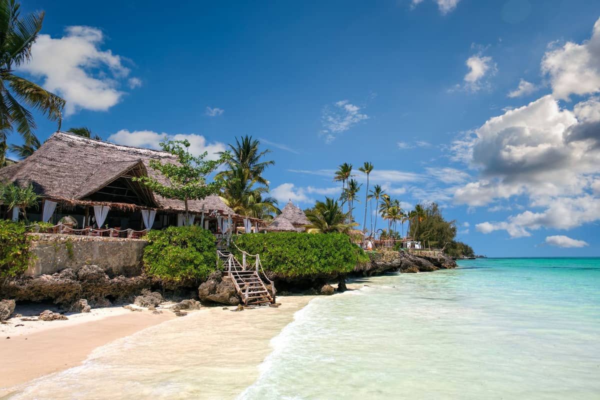 Strandresort in Sansibar mit Bungalows mit Strohdach, Palmen und türkisfarbenem Wasser unter einem sonnigen blauen Himmel.