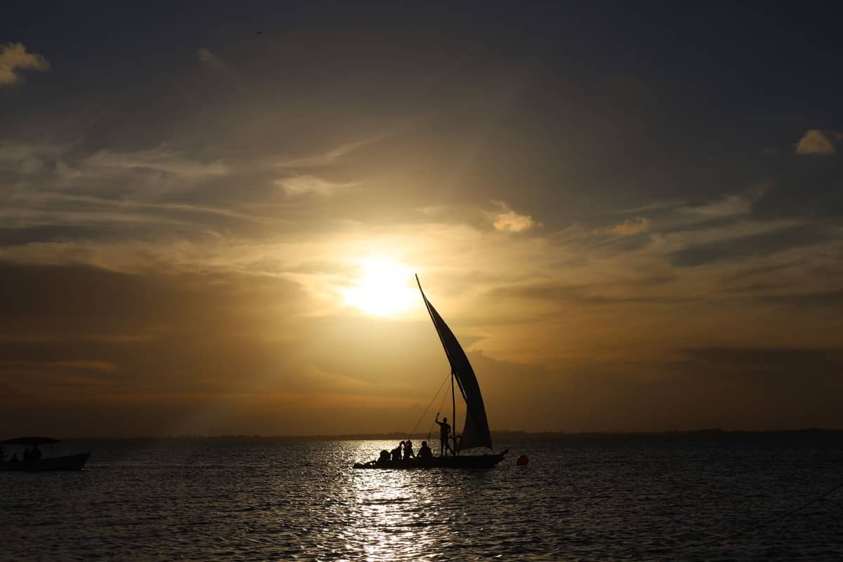 Silhouette einer kleinen Gruppe auf einem traditionellen Dhow-Segelboot bei Sonnenuntergang, mit goldenem Himmel und ruhigem Wasser, romantische Stimmung in Sansibar.