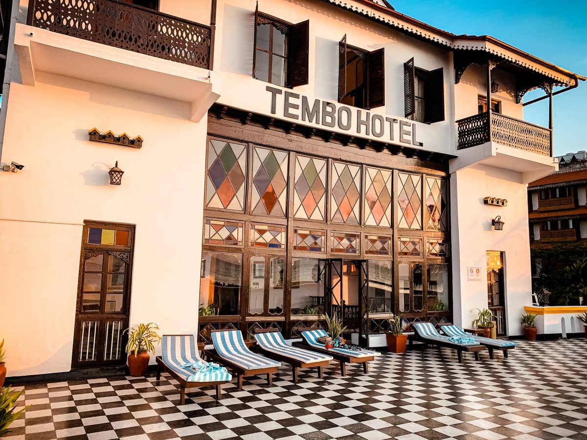 Fachada del Tembo Hotel en Stone Town, Zanzíbar, con tumbonas de rayas, suelo de cuadros y arquitectura tradicional bajo la luz del atardecer.