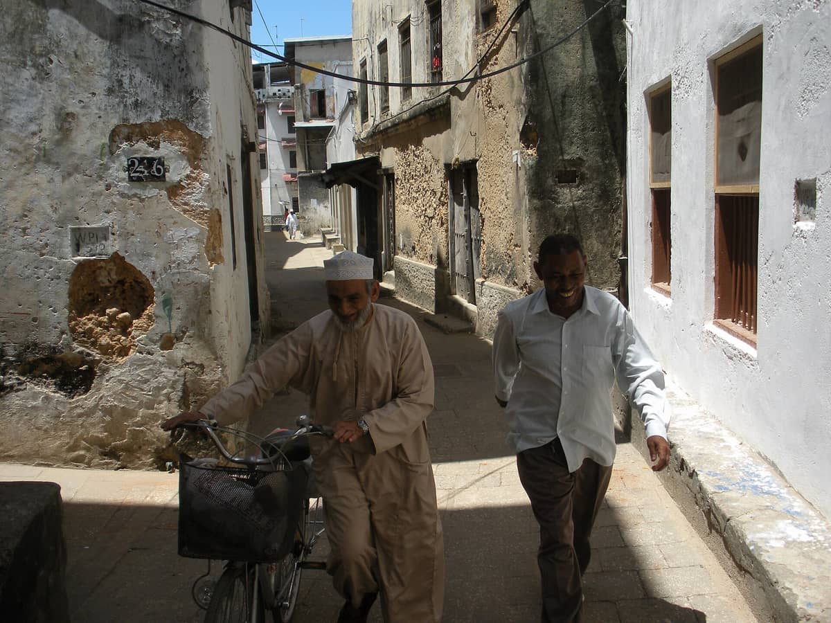 Dos hombres caminando y en bicicleta por un estrecho callejón soleado flanqueado por históricos edificios enlucidos en Stone Town, Zanzíbar.