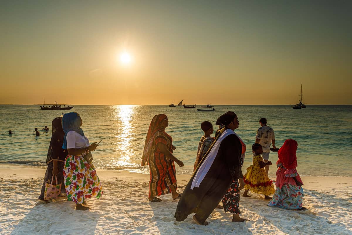 Miejscowi w kolorowych strojach spacerują po piaszczystej plaży na Zanzibarze o zachodzie słońca, w tle łodzie na wodzie i świąteczna atmosfera.