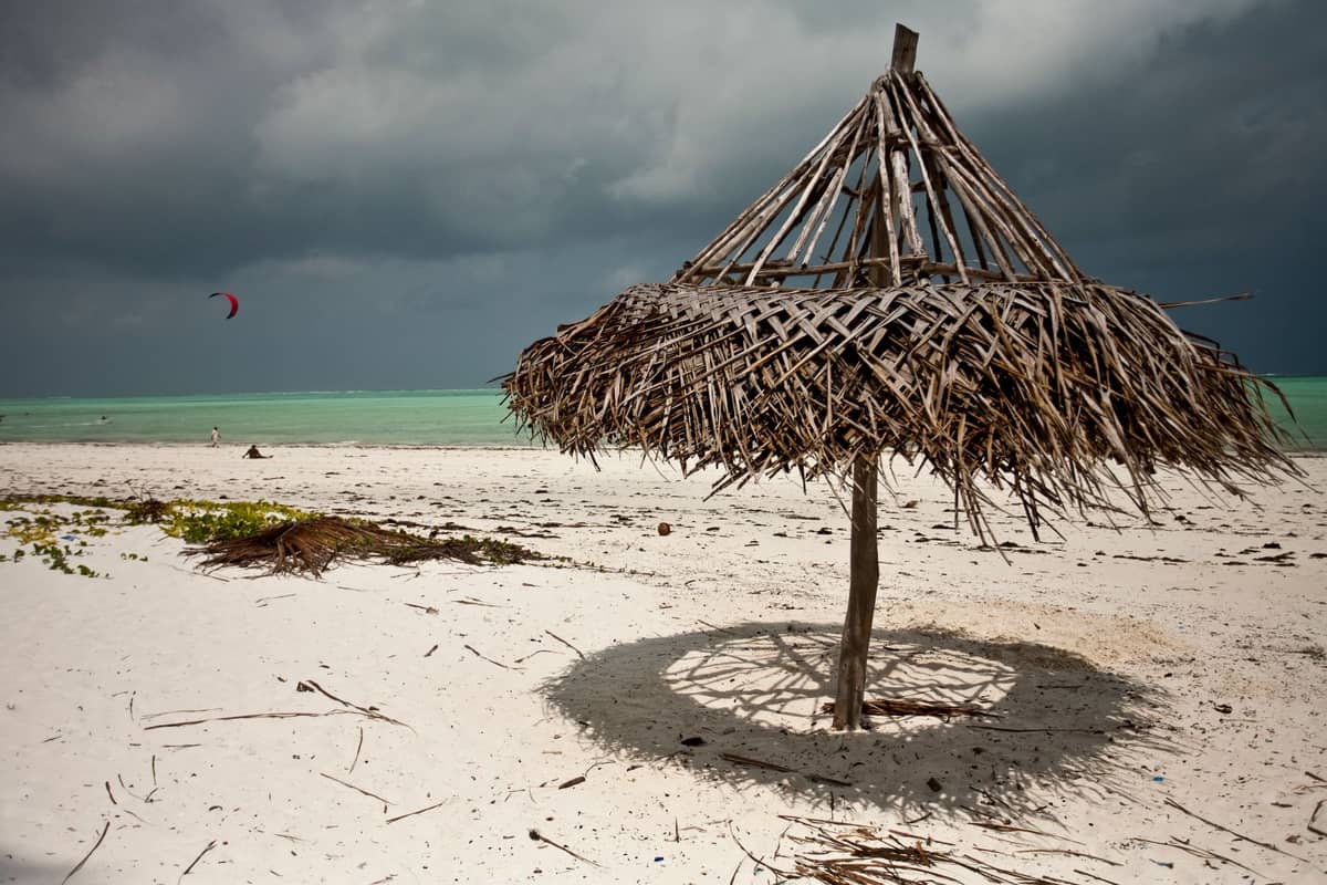 Thatched umbrella on a white sand beach under moody December skies and turquoise water, with distant kite surfer and seaweed visible.
