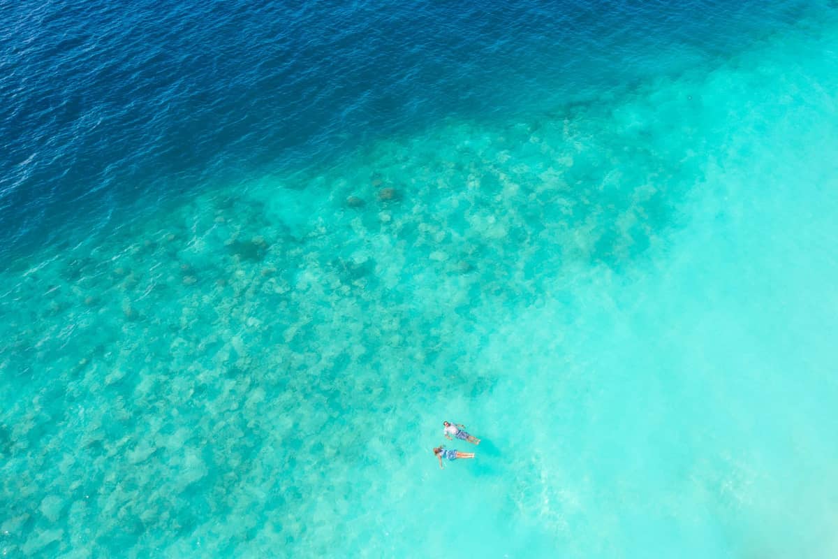Aerial view of two people snorkeling in clear turquoise water over a coral reef, with deep blue ocean nearby.