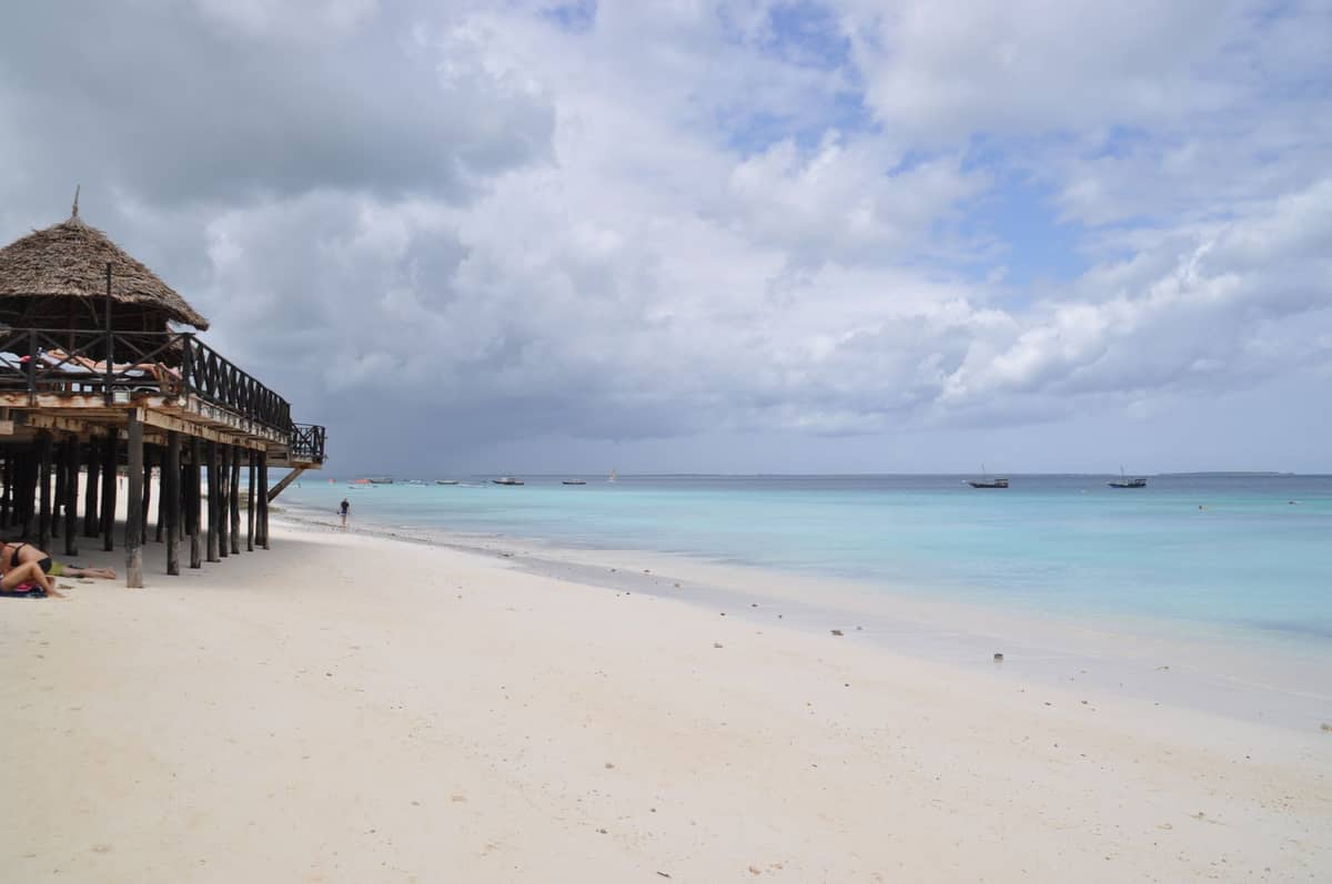 Szeroka piaszczysta plaża na Zanzibarze z spokojną turkusową wodą, wypoczywającymi i pływającymi ludźmi oraz łódkami kołyszącymi się na wodzie podczas delikatnego lipcowego zachodu słońca.