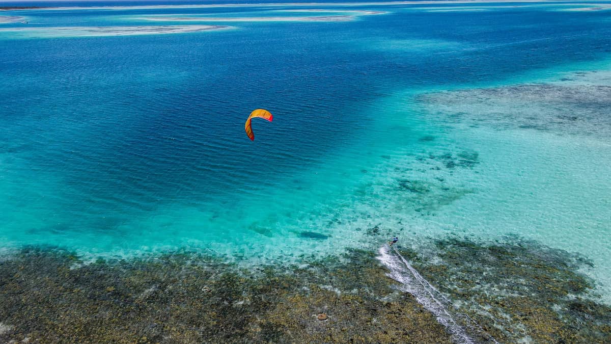 Luftaufnahme eines Kitesurfers, der über türkisfarbenes Wasser und ein Korallenriff nahe einem Sandstrand von Sansibar gleitet.