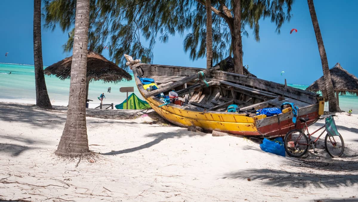 Plaża Paje na Zanzibarze: tradycyjny dhow na białym piasku, turkusowa woda laguny, palmy i kitesurferzy w oddali.