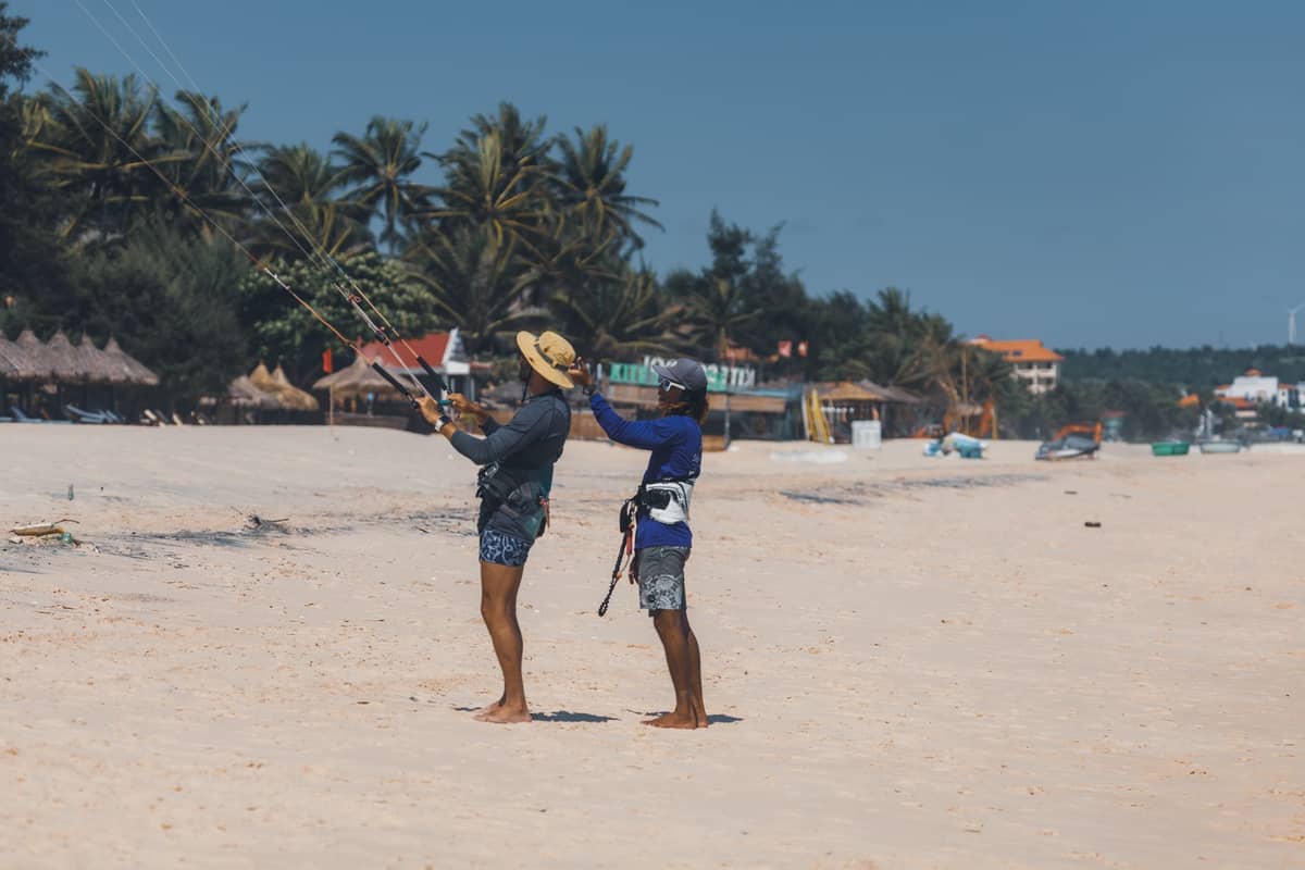 Zwei Personen stehen an einem tropischen Sandstrand, wobei eine der anderen beibringt, wie man eine Kitesurfbar handhabt, mit Palmen und Strandhütten im Hintergrund.
