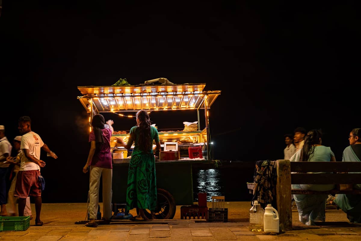 แผงขายอาหารริมถนนยามค่ำคืนที่ประดับด้วยไฟประดับ ผู้คนกำลังสั่งอาหารและนั่งบนม้านั่งริมน้ำใน Stone Town