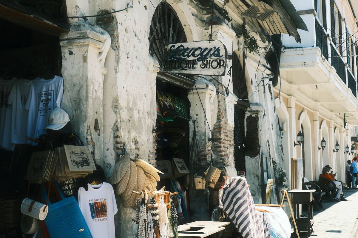 ถนนใน Stone Town พร้อมร้านของที่ระลึกสไตล์รัสติกที่จำหน่ายงานหัตถกรรม ผ้า และตะกร้าใต้ป้ายเก่า มีชาวท้องถิ่นและนักท่องเที่ยวเดินอยู่ใกล้เคียง