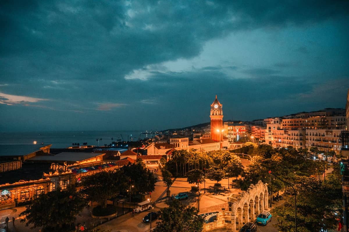 Vista nocturna sobre Stone Town en Zanzíbar con torre del reloj iluminada, farolas encendidas, océano Índico y edificios históricos bajo un cielo de tonos azules.