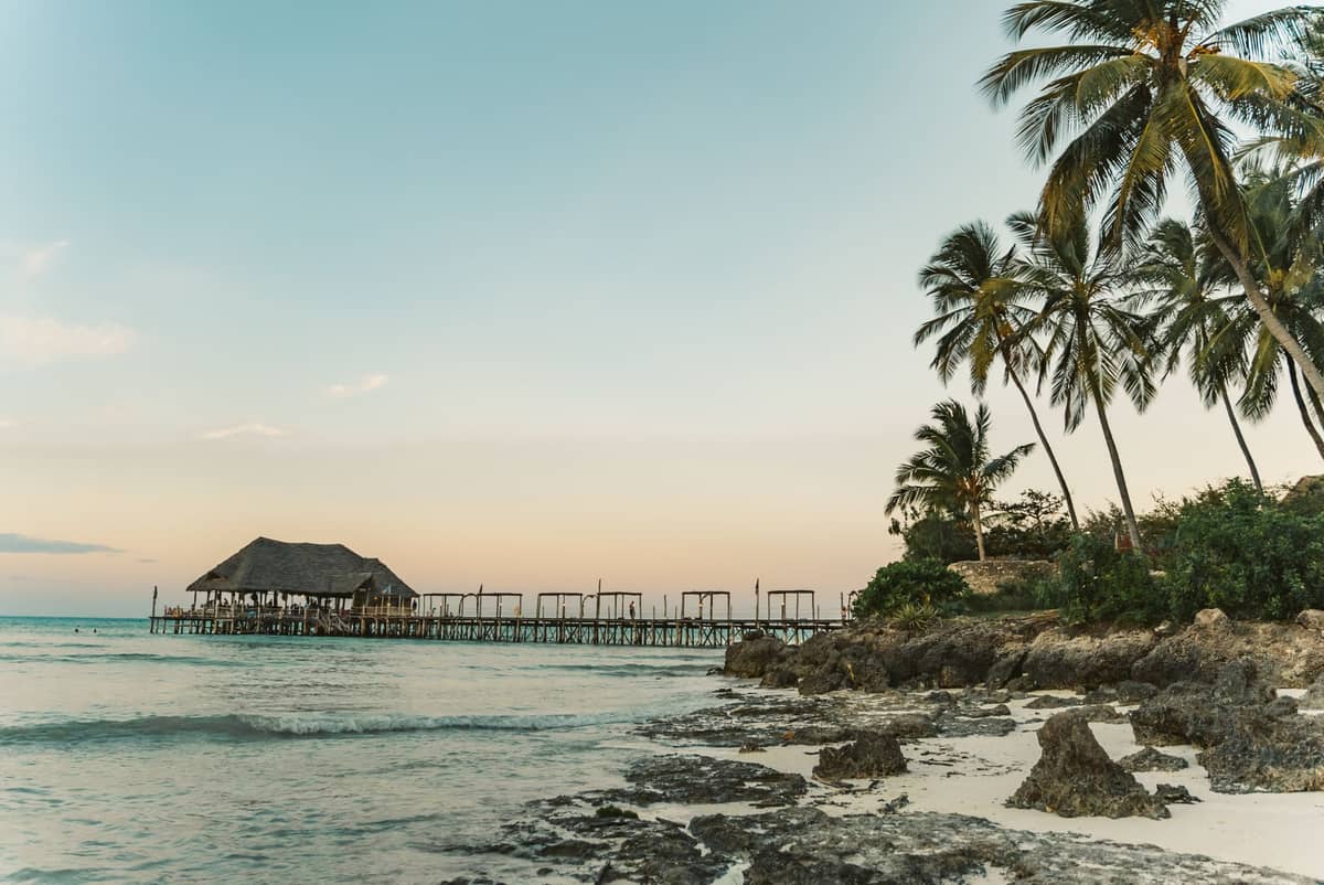 Playa tropical al atardecer con palmeras, orilla rocosa y un muelle de madera que se extiende sobre el agua en Zanzíbar.