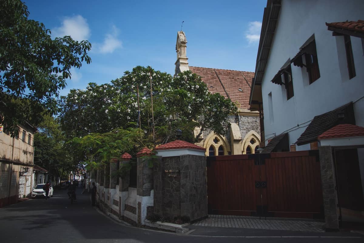 Street view of Christ Church Cathedral in Stone Town, Zanzibar, partially visible behind trees and a wall with blue sky above.