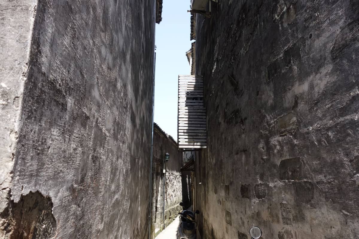 Narrow alleyway with high stone walls and a wooden balcony connecting buildings in bright sunlight.