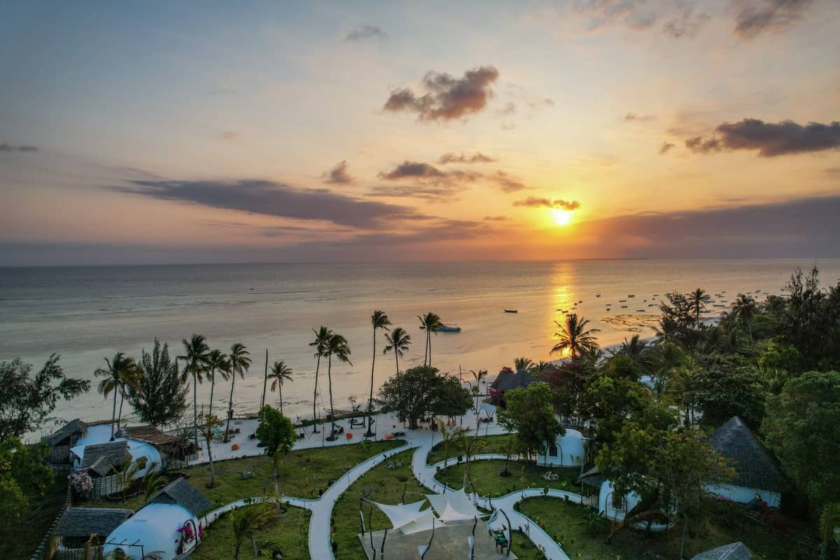 Luftaufnahme eines Strandes und Resortgartens in Sansibar bei Sonnenuntergang mit Palmen und Booten entlang der Küste.