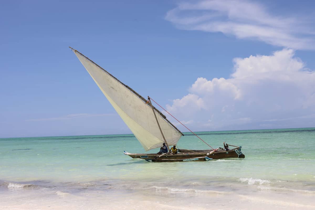 Traditionelles hölzernes Dhow mit einem großen Segel auf türkisfarbenem Wasser nahe einem weißen Sandstrand und blauem Himmel in Sansibar.