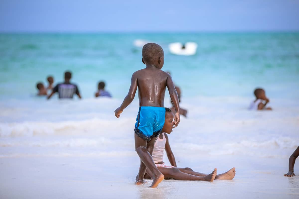 Dzieci bawiące się i pływające w płytkiej turkusowej wodzie na piaszczystej plaży na Zanzibarze, w tle czysta woda i ludzie.