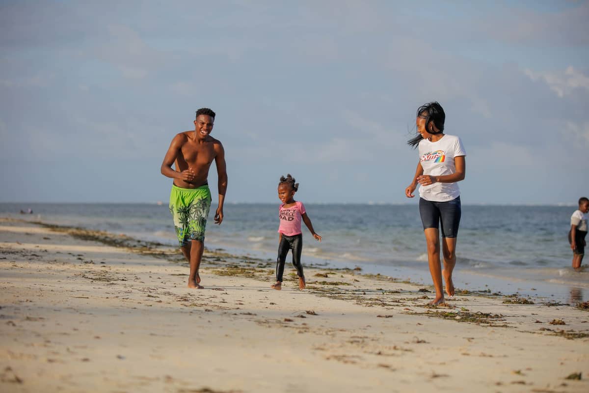 Drei Kinder rennen und spielen zusammen an einem Sandstrand mit dem Ozean im Hintergrund unter sonnigem Himmel.
