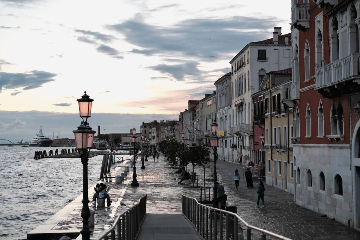 Blick auf die Zattere-Promenade in Venedig bei Sonnenuntergang: Menschen schlendern entlang des breiten Ufers am Giudecca-Kanal, gesäumt von traditionellen Laternen.