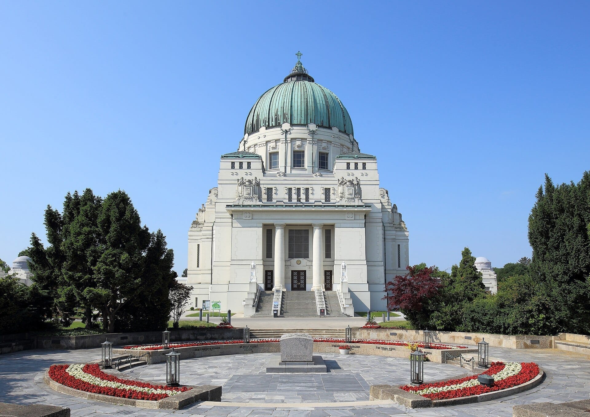 Frontansicht der Karl-Borromäus-Kirche am Zentralfriedhof Wien an einem sonnigen Tag, mit bunten Blumenbeeten und gepflegtem Grün.