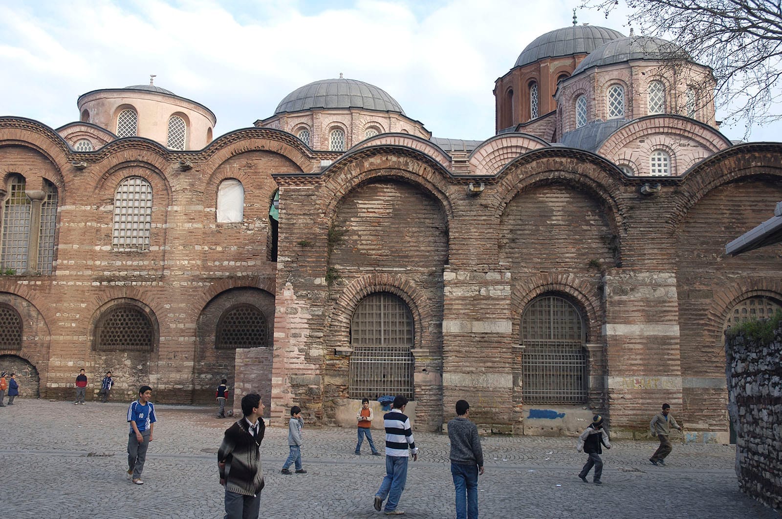 Vista esterna delle mura in mattoni e delle cupole della Moschea Zeyrek, con bambini che giocano nel cortile acciottolato in una giornata luminosa.